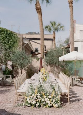 An elegant outdoor dining setup with a long table, chairs, and floral arrangements, situated in a courtyard with palm trees and surrounding greenery.