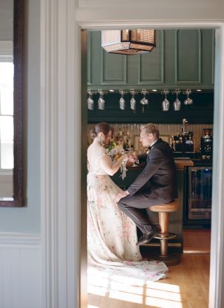 A couple is sitting in a cozy kitchen, possibly celebrating with drinks. The woman is in a gown and the man is in a suit, suggesting a formal occasion.