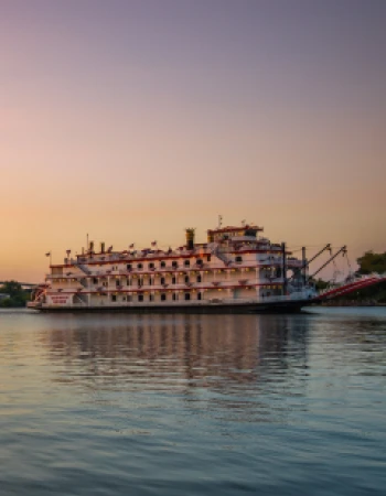 A large riverboat with multiple decks is on a calm body of water at sunset with a pastel-colored sky in the background.