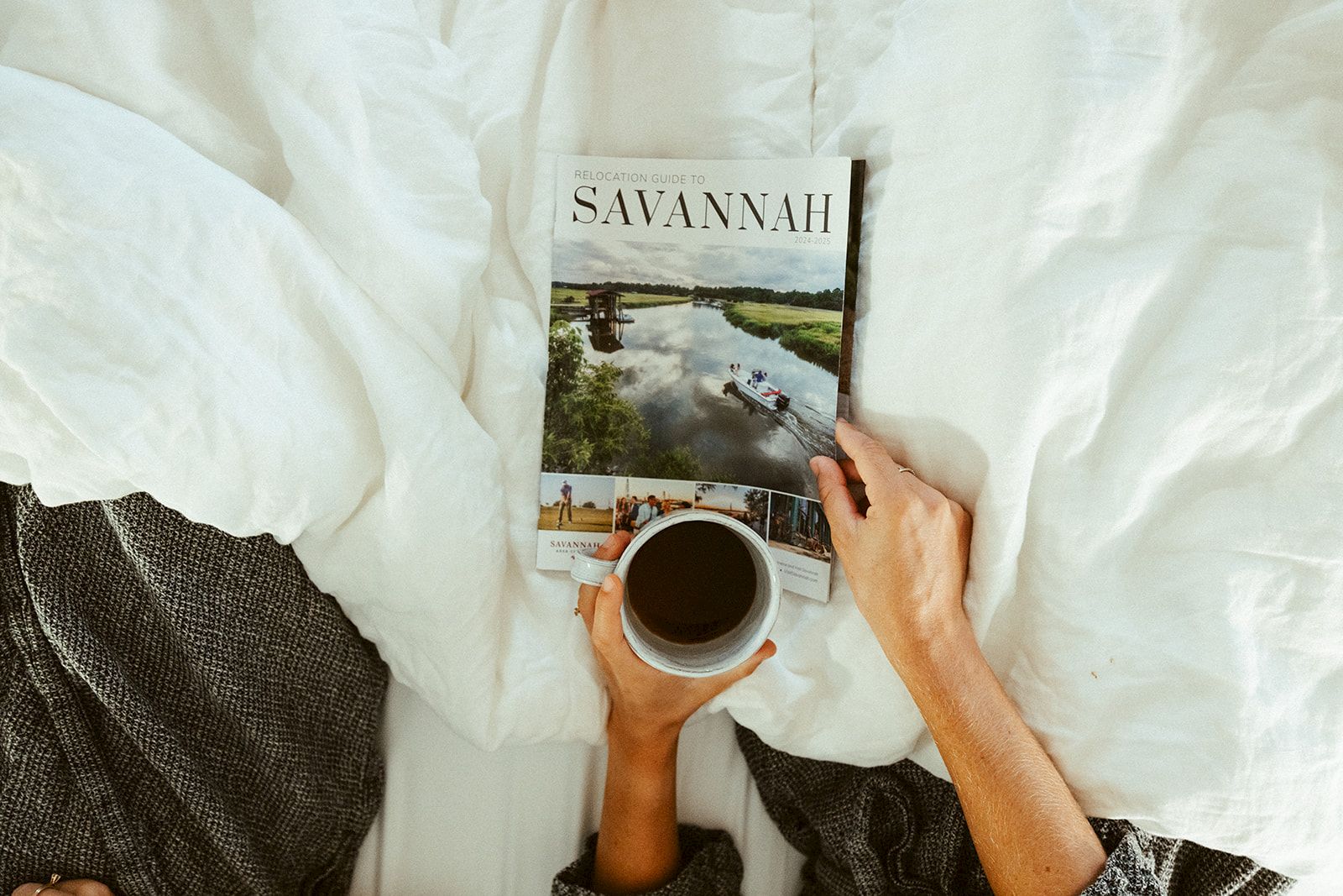 A person sits in bed reading a magazine titled &ldquo;SAVANNAH&rdquo; while holding a cup of coffee, with a cozy white duvet and blanket nearby.