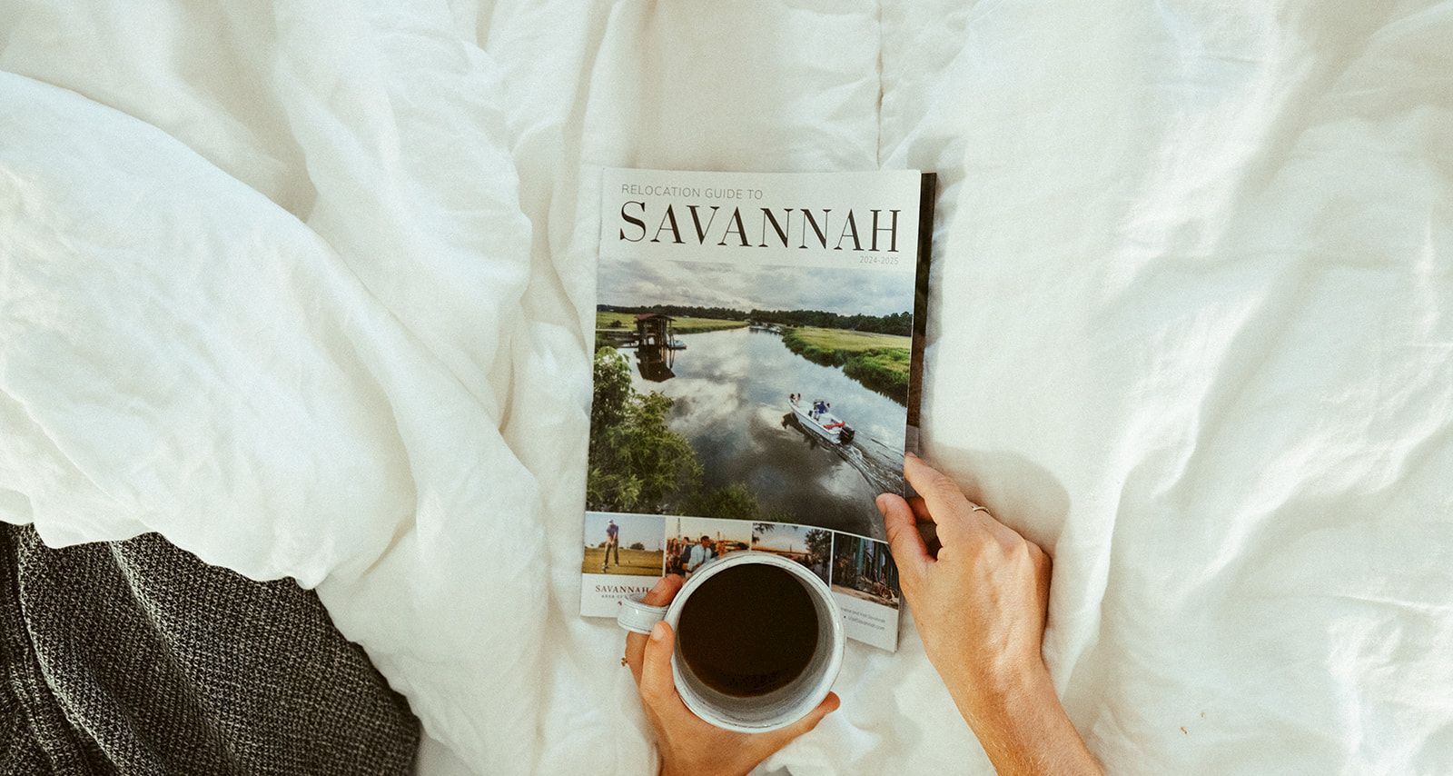 A person sits in bed reading a magazine titled &ldquo;SAVANNAH&rdquo; while holding a cup of coffee, with a cozy white duvet and blanket nearby.