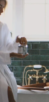 A woman in a white robe is seated by a bathtub, holding a jar in a bathroom with green tiles and a window in the background.