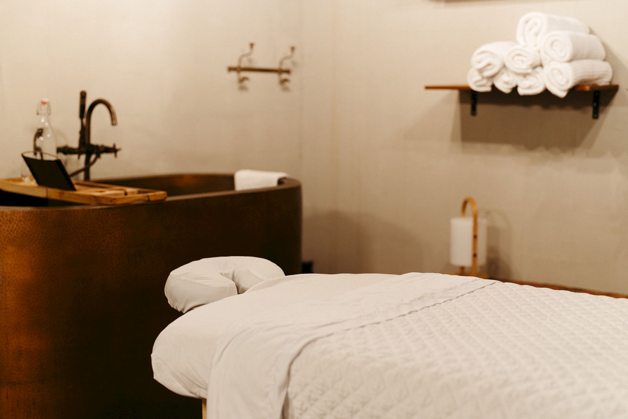 A spa room with a massage table, stacked towels on a shelf, and a nearby tub with a faucet, creating a relaxing atmosphere.