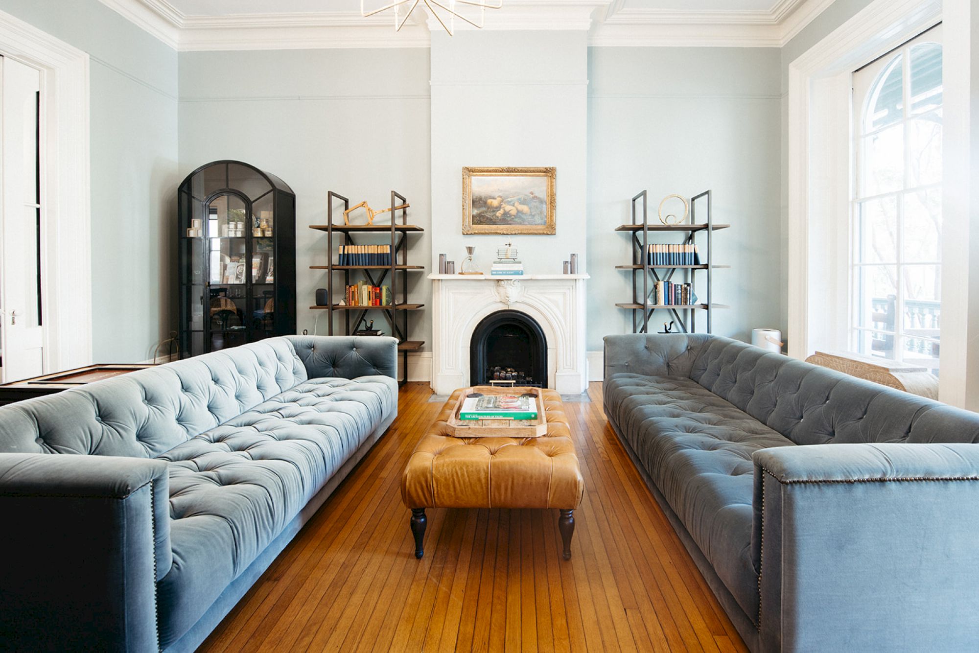 A stylish living room with two blue sofas, a central ottoman, wooden floor, fireplace, bookshelves, and a framed painting on the wall.