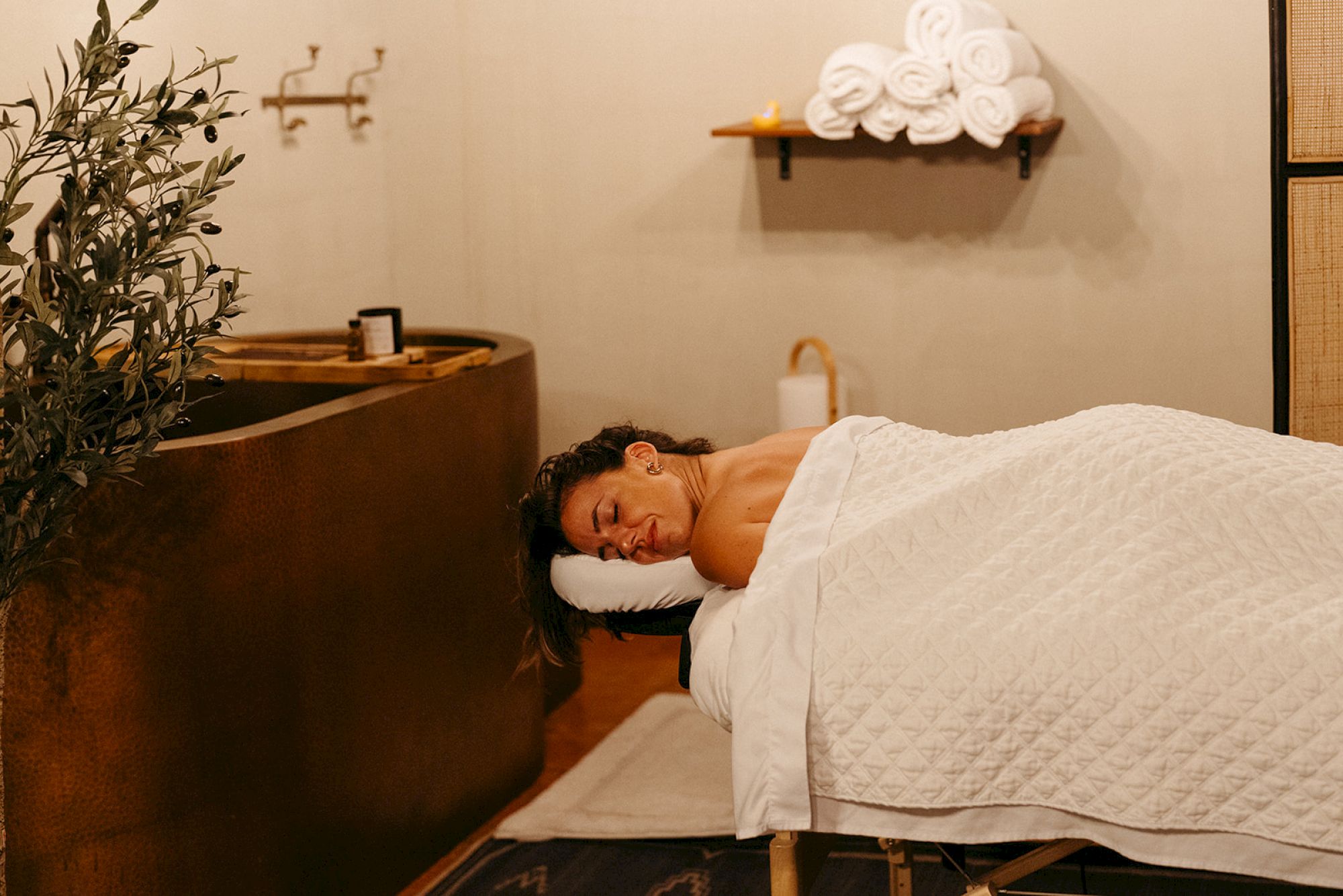 A person is lying on a massage table under a white blanket in a spa setting, with towels neatly stacked on a shelf.