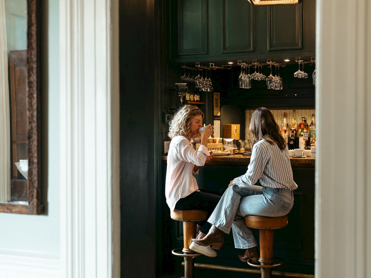 Two people sit on bar stools engaged in conversation at a cozy bar, with glasses and bottles seen in the background.