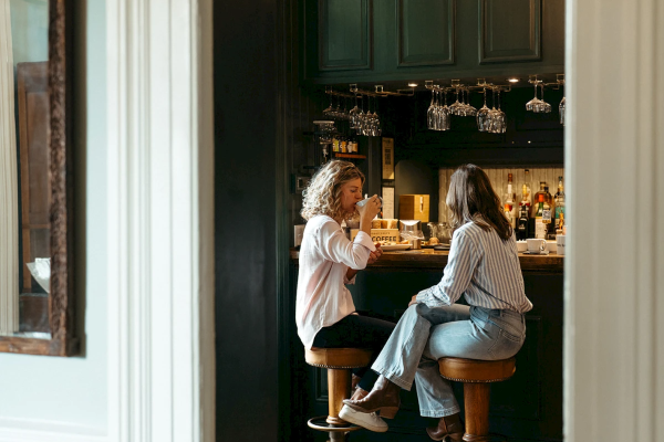 Two people sitting at a bar counter, engaged in conversation, with drinks and bottles visible behind them.