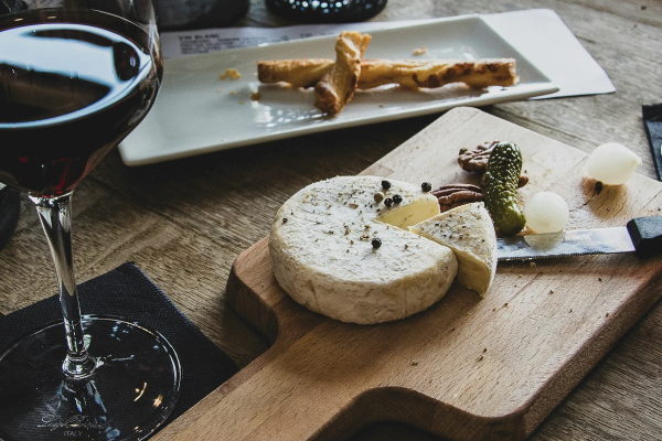 A glass of red wine with cheese, crackers, pickles, and onions on a wooden board, placed on a textured table surface.