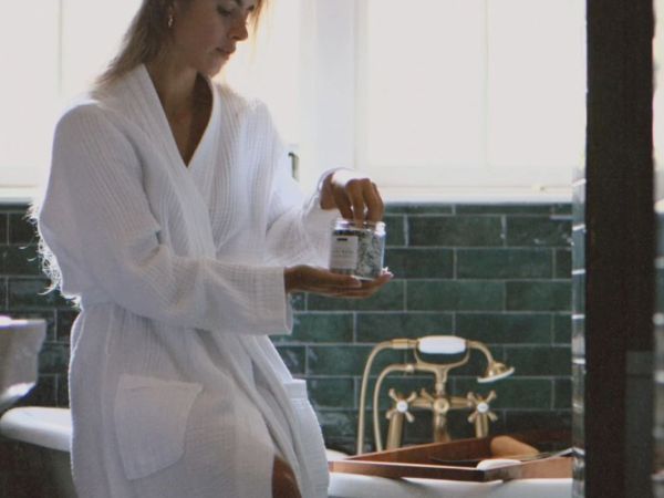 A woman in a white robe pours from a small container at a sink with a brass faucet against green tile, in a bathroom.