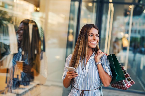 A woman is happily shopping, holding a phone and carrying shopping bags, standing near a glass storefront.