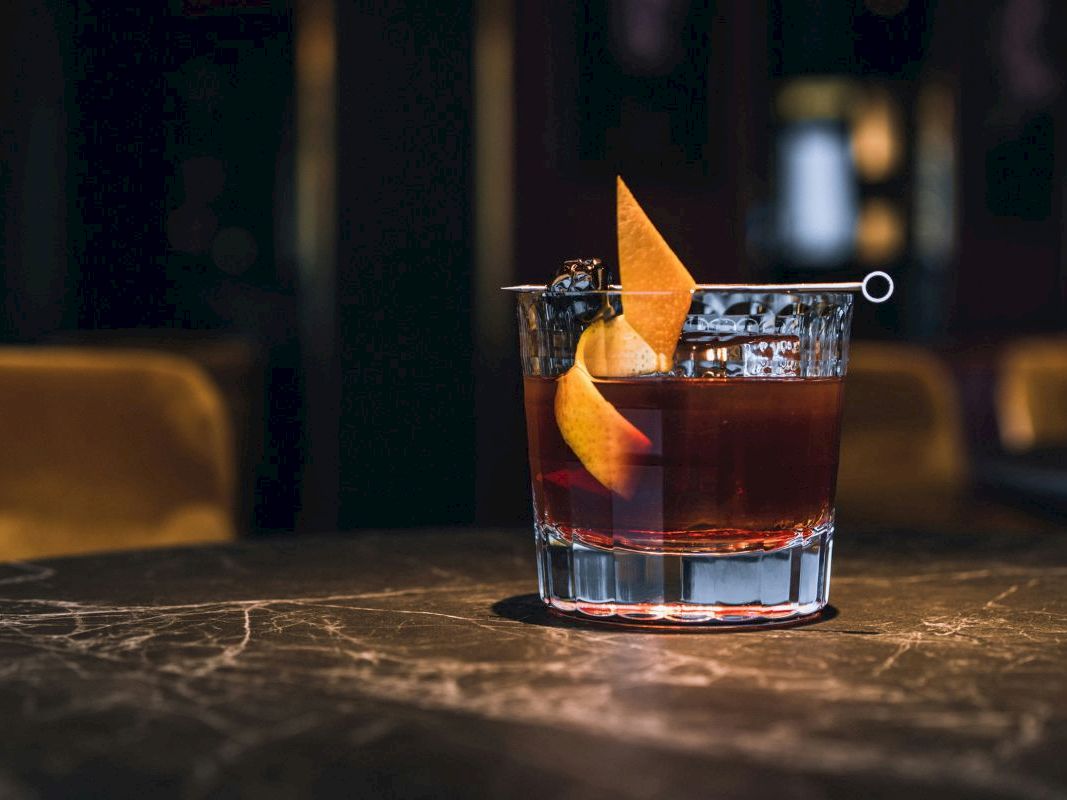 A rocks glass cocktail with amber liquor, ice, and an orange peel garnish on a bar counter.