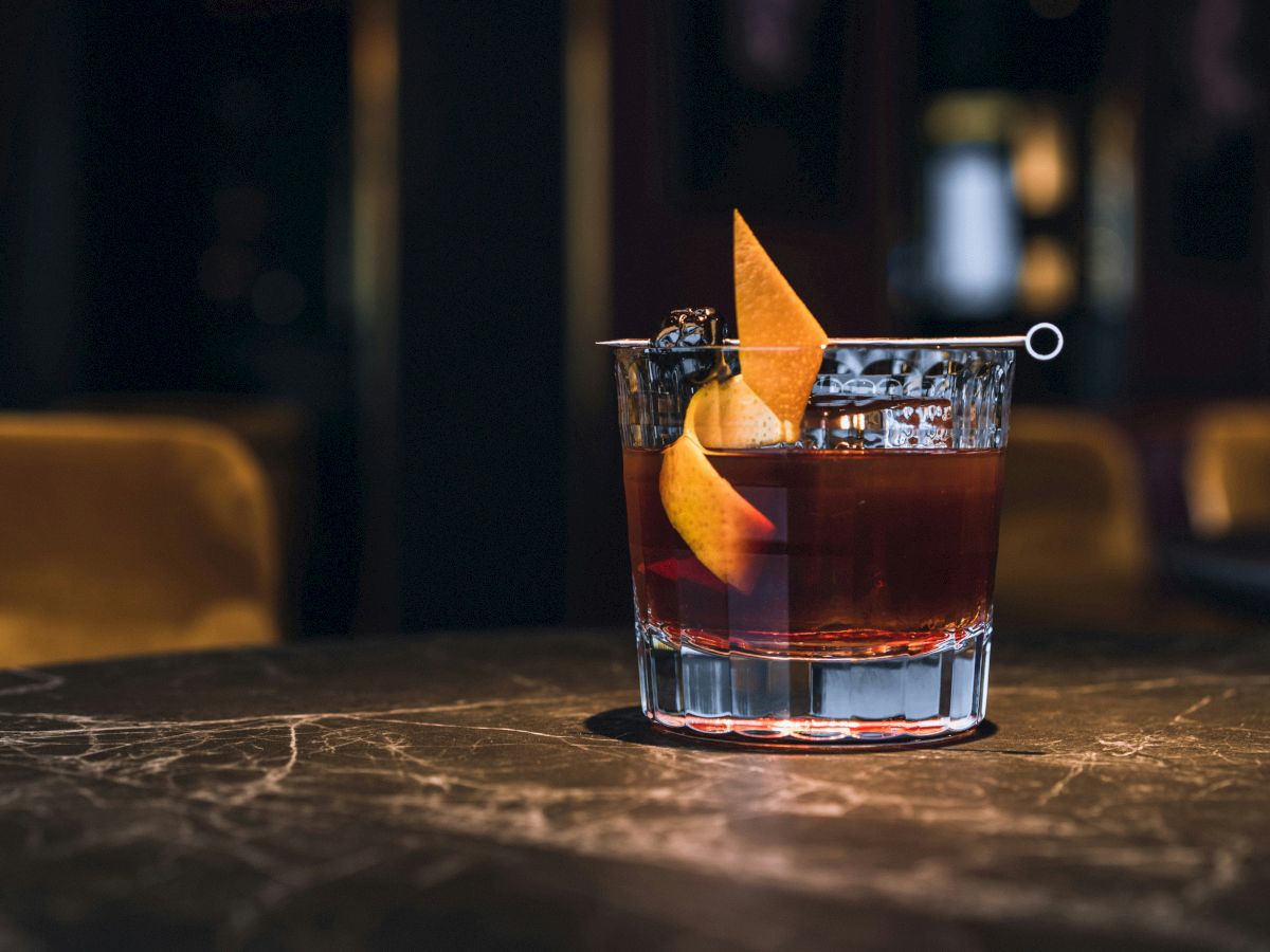 A cocktail in a glass garnished with an orange peel sits on a dark marble table in a dimly lit setting.