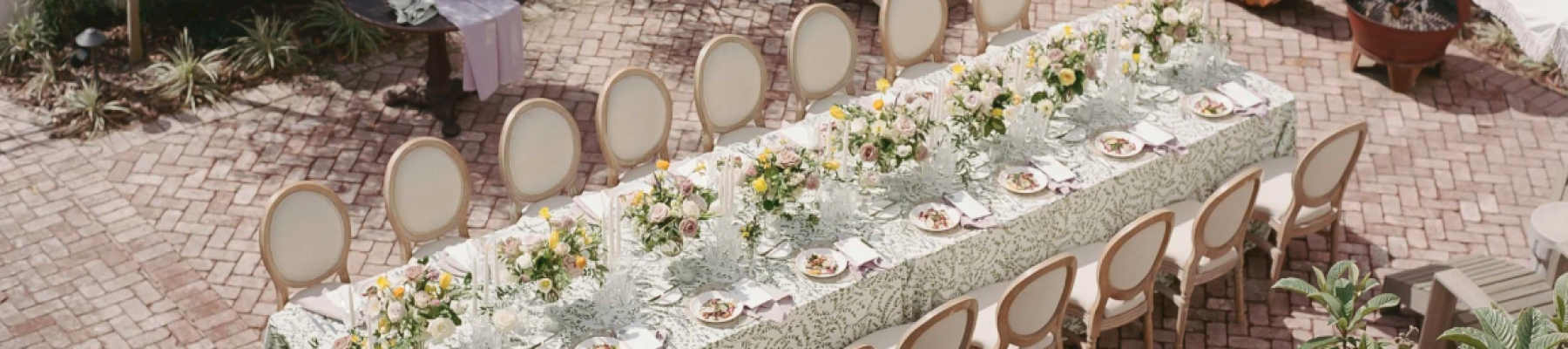 An outdoor table is elegantly set for a gathering, adorned with flowers and surrounded by twelve chairs on a brick patio.