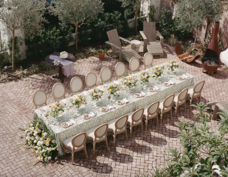 An outdoor table is elegantly set for a gathering, adorned with flowers and surrounded by twelve chairs on a brick patio.