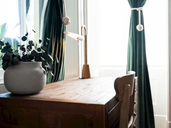 A wooden desk with a potted plant and a lamp stands by a window with green curtains.