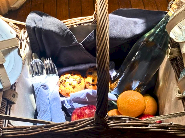 A picnic basket with bread, cookies, oranges, a bottle of wine, napkins, and utensils lined up inside the basket.