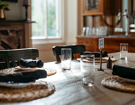 A dining table is set with woven placemats, glasses, folded black napkins, and a salt shaker in a warmly-lit room.