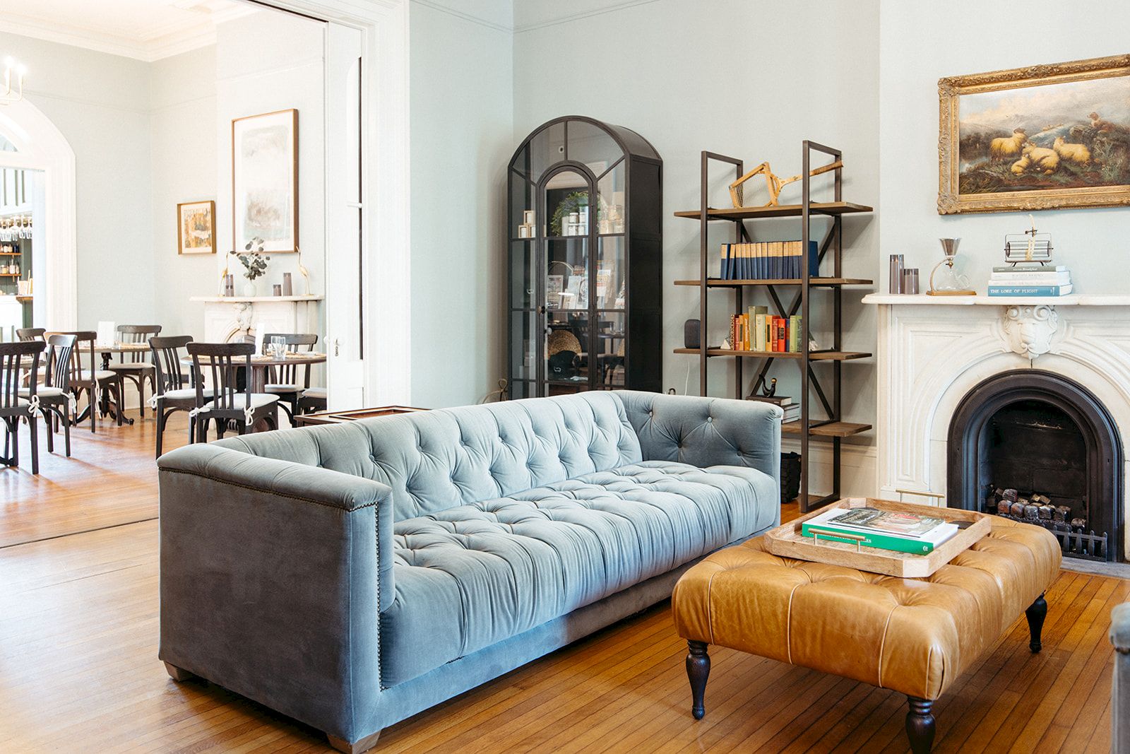 A stylish living room with a blue tufted sofa, leather ottoman, wooden floor, fireplace, and shelves; bright dining area in the background.