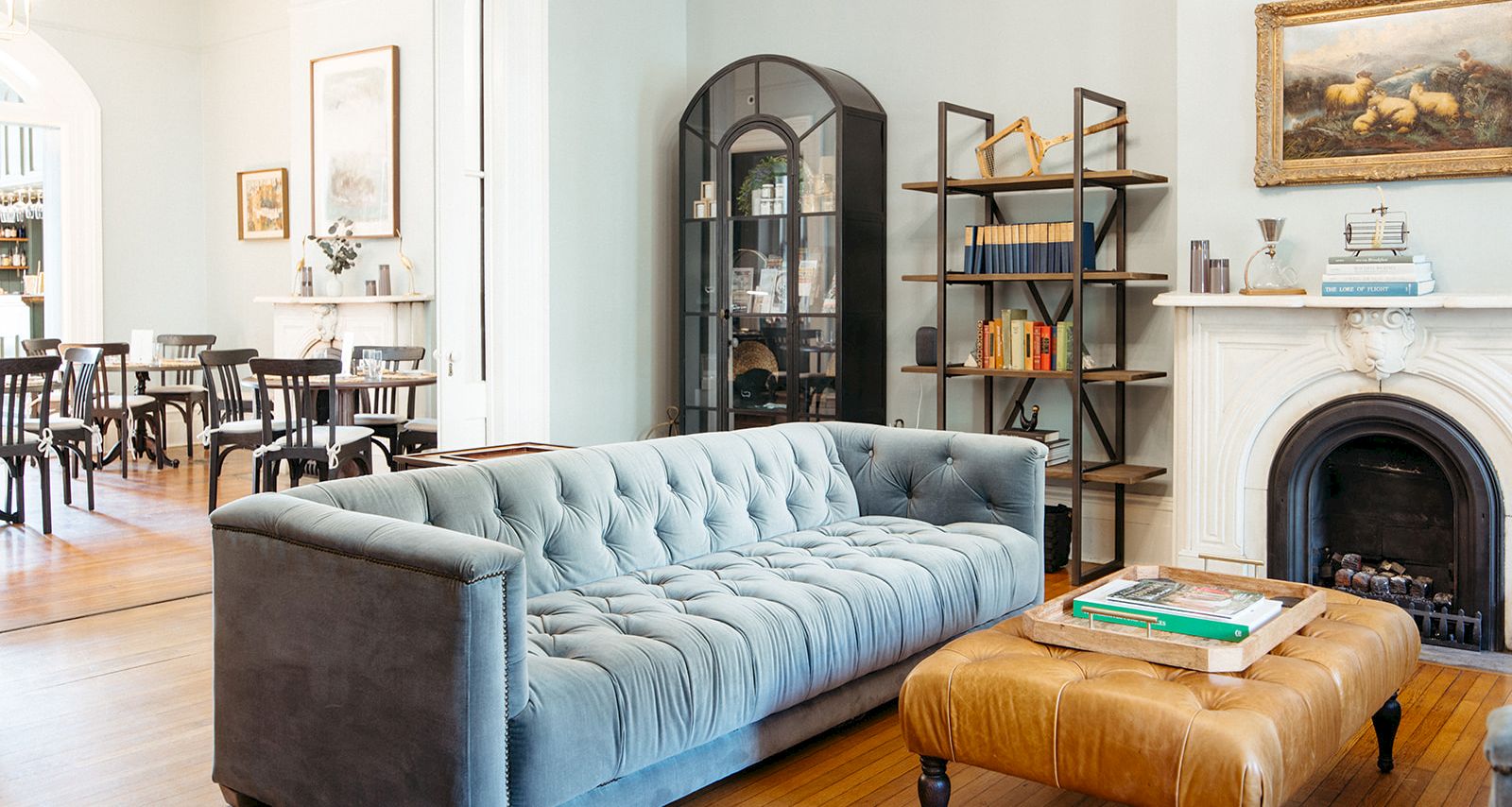A stylish living room with a blue tufted sofa, leather ottoman, wooden floor, fireplace, and shelves; bright dining area in the background.