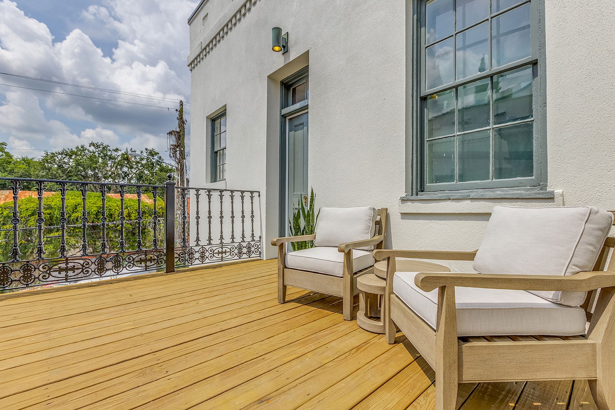 A sunny wooden deck with two cream-cushioned chairs, a small side table, potted plant, and a white building with large windows.