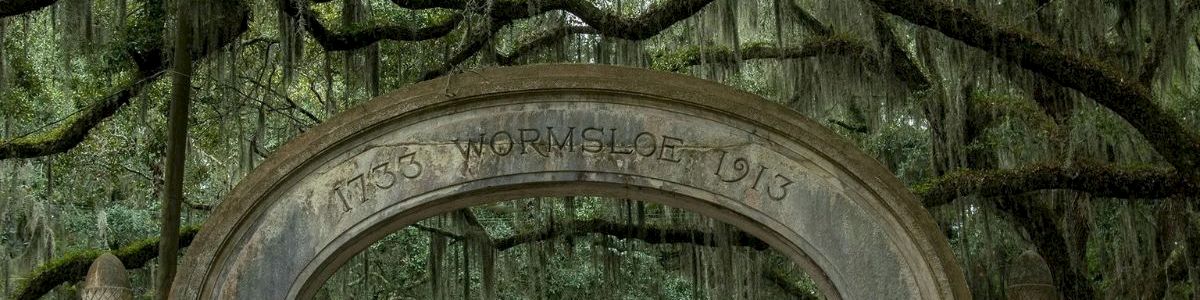 A moss-covered arch entrance stands in a steamy oak grove draped with Spanish moss, with &ldquo;Wormsloe&rdquo; carved on top.