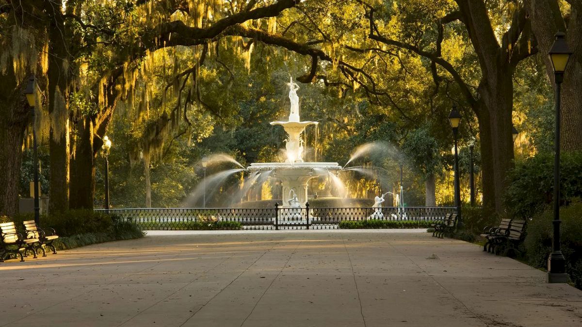 A grand fountain sprays water in a shaded park, draped with Spanish moss, with benches and lamps along the path.