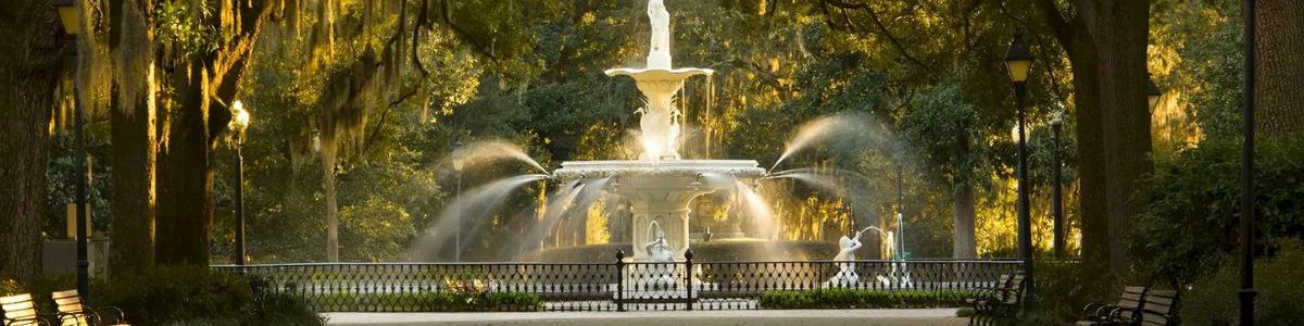 A grand fountain sprays water in a shaded park, draped with Spanish moss, with benches and lamps along the path.