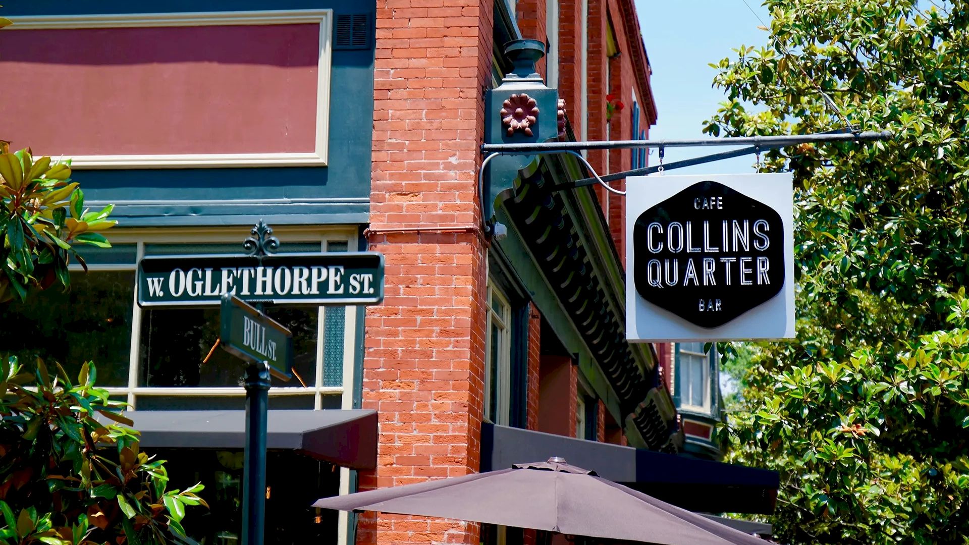 A brick building on a street corner with a &ldquo;Collins Quarter&rdquo; sign, green trim, balcony, trees, and a large umbrella in front, sunny day.