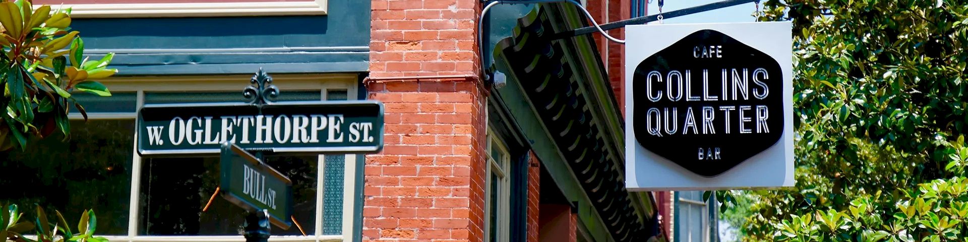 A brick building on a street corner with a &ldquo;Collins Quarter&rdquo; sign, green trim, balcony, trees, and a large umbrella in front, sunny day.