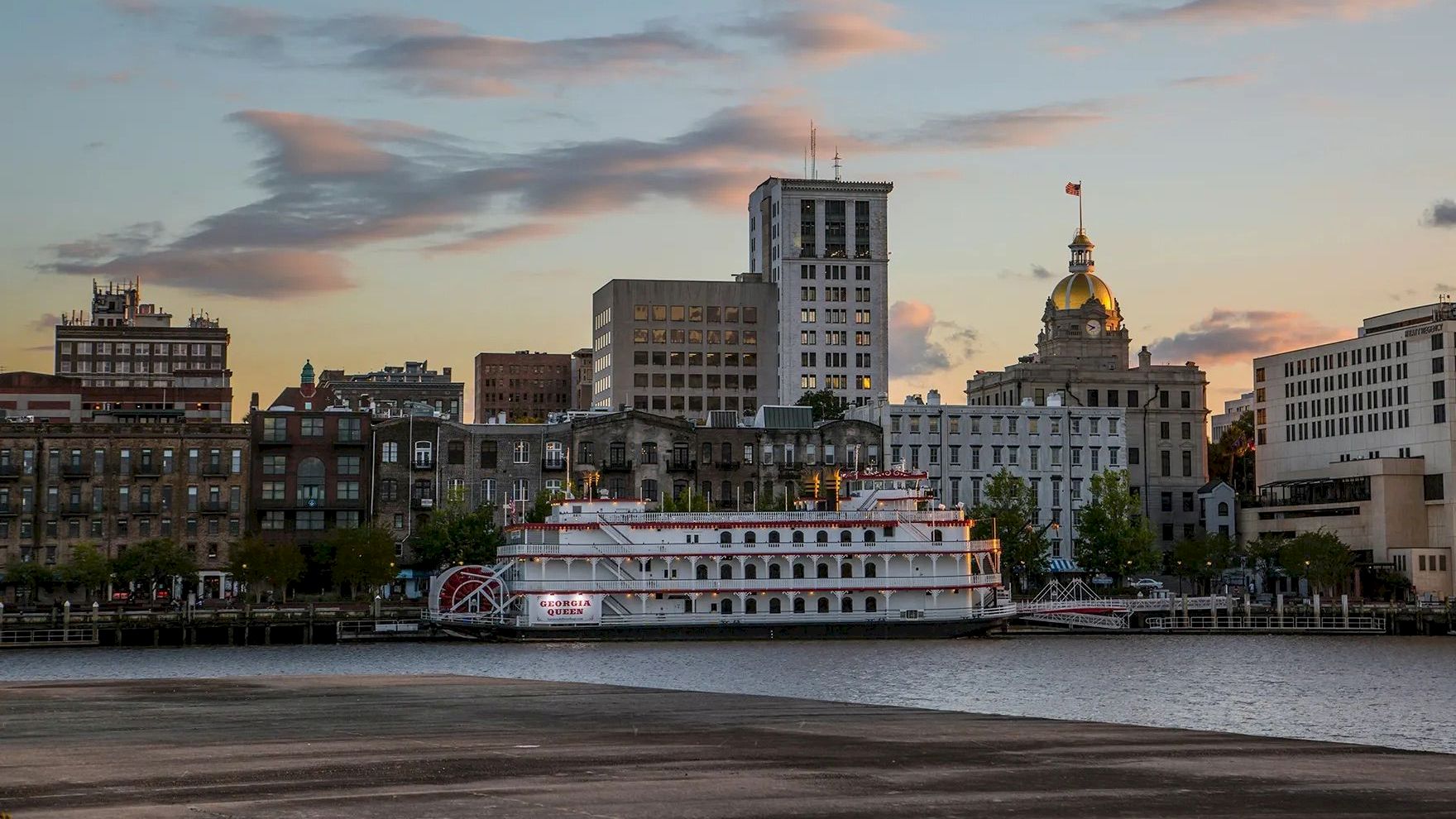 A city waterfront at dusk with a river, a white passenger boat docked by the quay, and skyline buildings glowing softly as clouds drift above.