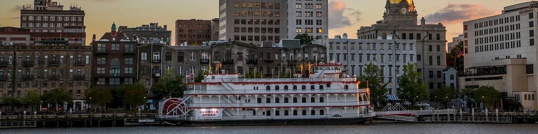 A city waterfront at dusk with a river, a white passenger boat docked by the quay, and skyline buildings glowing softly as clouds drift above.