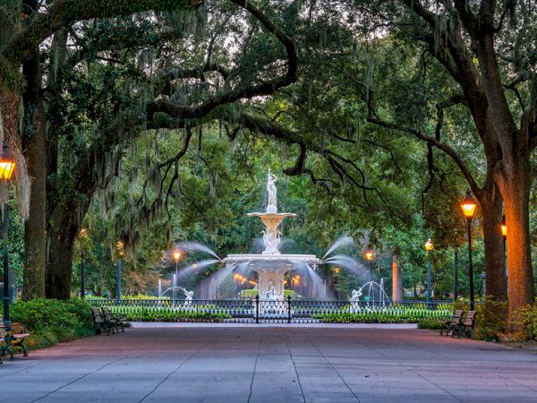 A serene park scene featuring a central fountain, surrounded by lush trees, lampposts, benches, and a tranquil pathway at dusk.