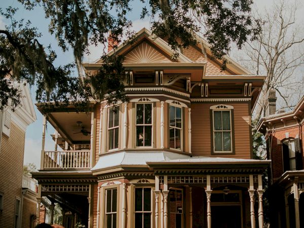 A tall, ornate two-story Victorian house with beige siding, bay windows, decorative trim, a wrapped porch, and trees framing the facade.
