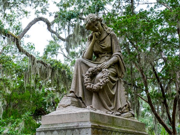 A statue of a seated grieving woman holding a flower wreath, set among green trees in a tranquil garden.