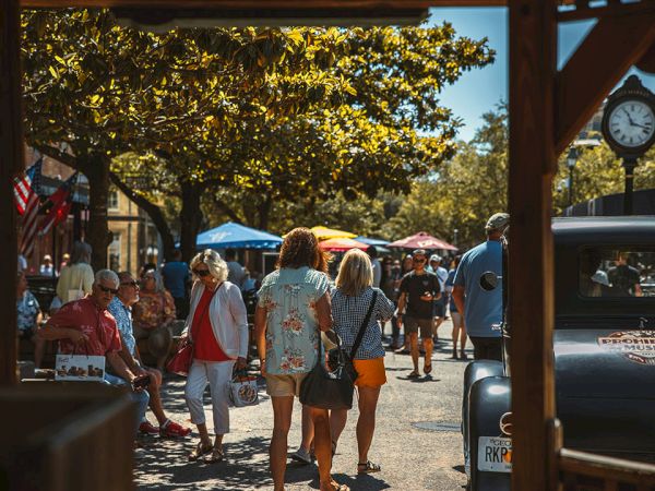 A sunny market scene with people strolling between stalls, tents, and trees; a clock tower and shops frame the busy, casual outdoor outing.