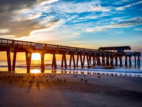 A wooden pier stretches into the ocean at sunset, casting long shadows on the wet sand as waves roll in.