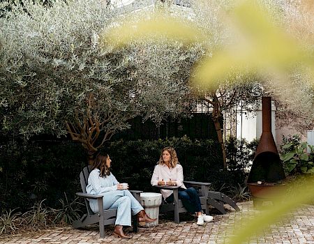 Two people sit in garden chairs, chatting beside a tree and brick oven, in a peaceful outdoor setting.