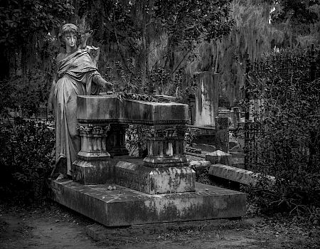 A weathered cemetery statue stands beside a stone tomb, draped in shadows and overgrown foliage, evokes quiet mourning and age.