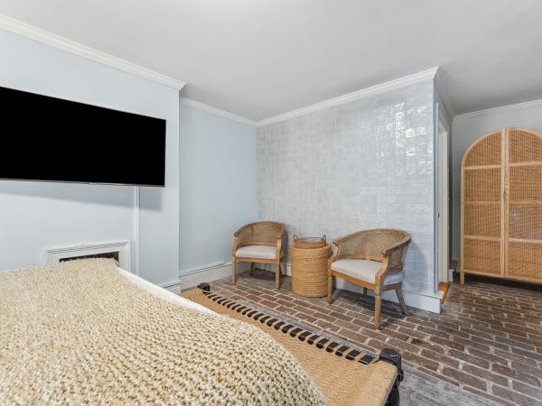 A cozy hotel room with a bed, a wall-mounted TV, two wicker chairs around a small table, a tiled wall, brick floor, and a wooden armoire, all softly lit.