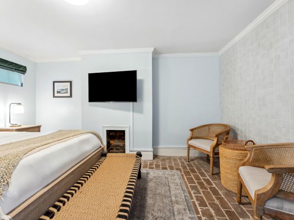 A hotel room with a bed, wall-mounted TV, fireplace nook, wicker chairs, and brick floor; a bench at the foot of the bed, blue walls, and a window.