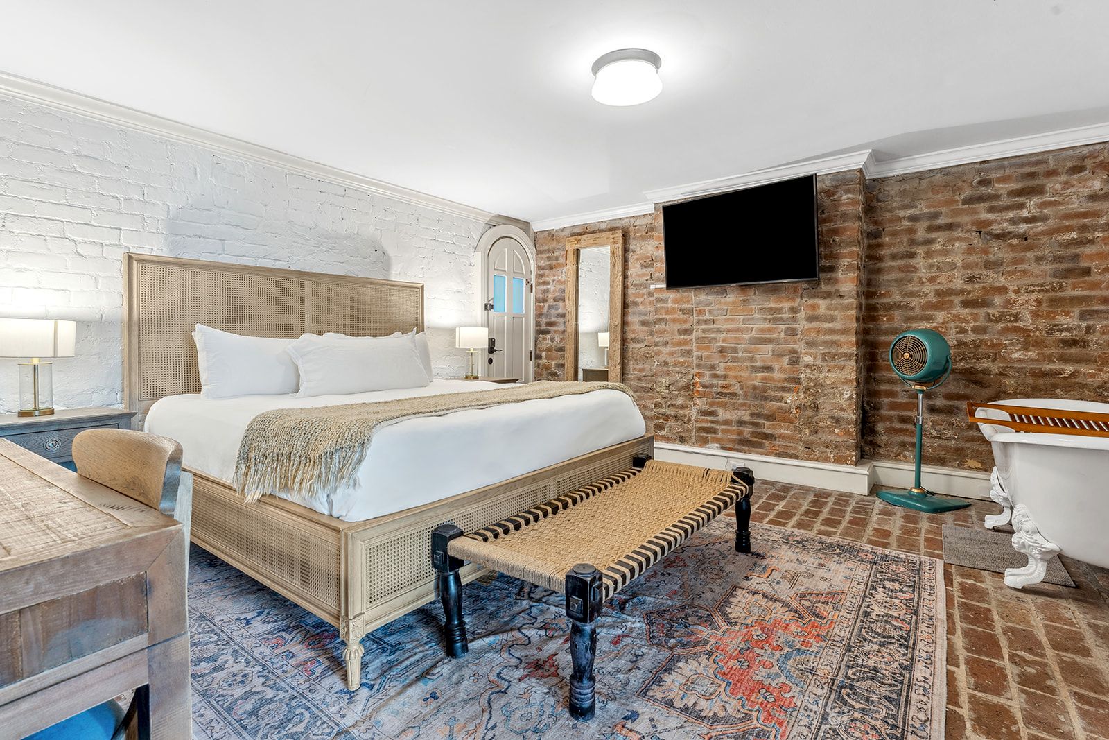 A stylish hotel room with a wooden bed, bench at foot, brick accent wall, mounted TV, vintage details, and a blue-and-rust rug&mdash;luxurious and cozy.