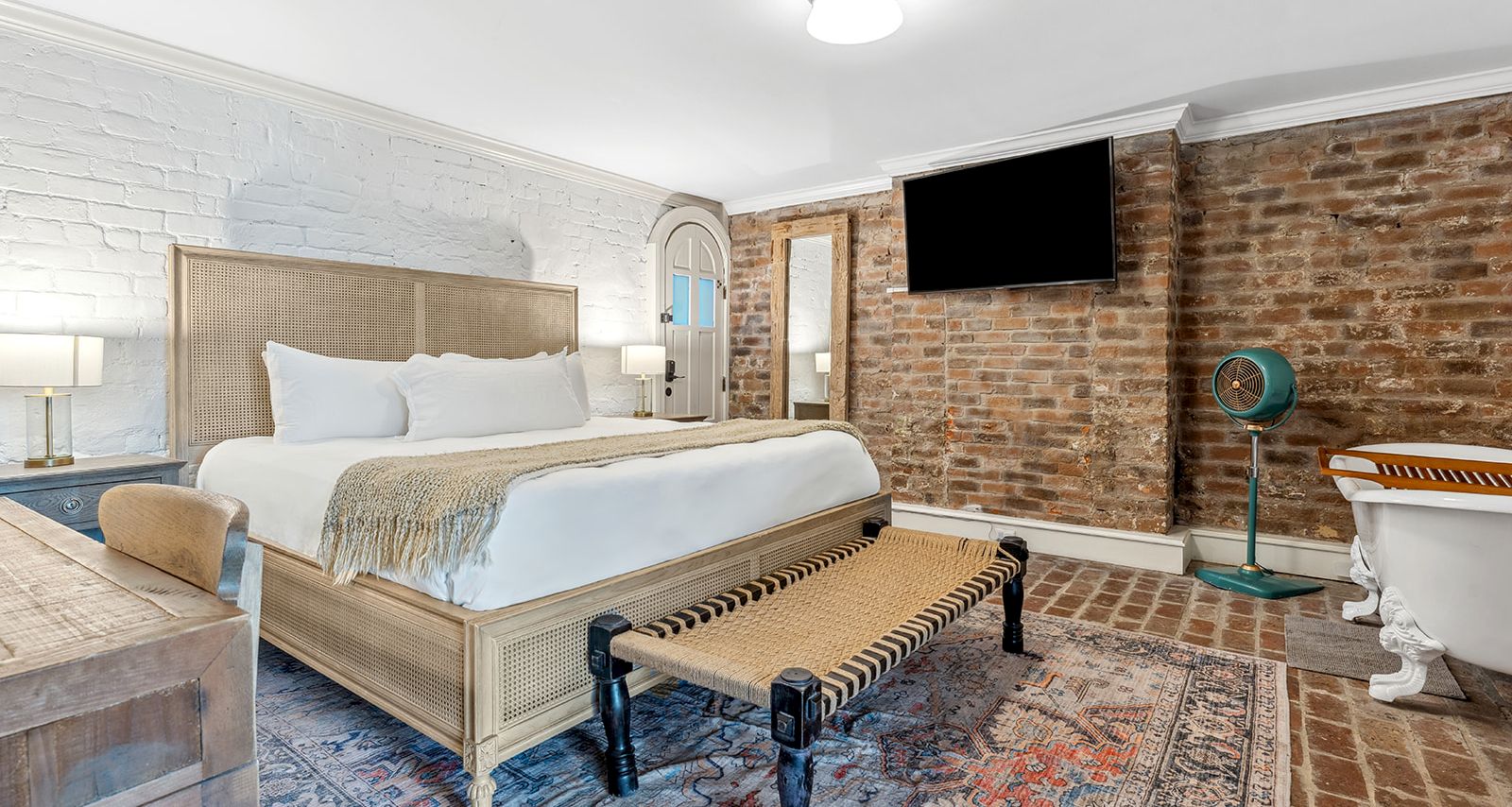 A stylish hotel room with a wooden bed, bench at foot, brick accent wall, mounted TV, vintage details, and a blue-and-rust rug&mdash;luxurious and cozy.