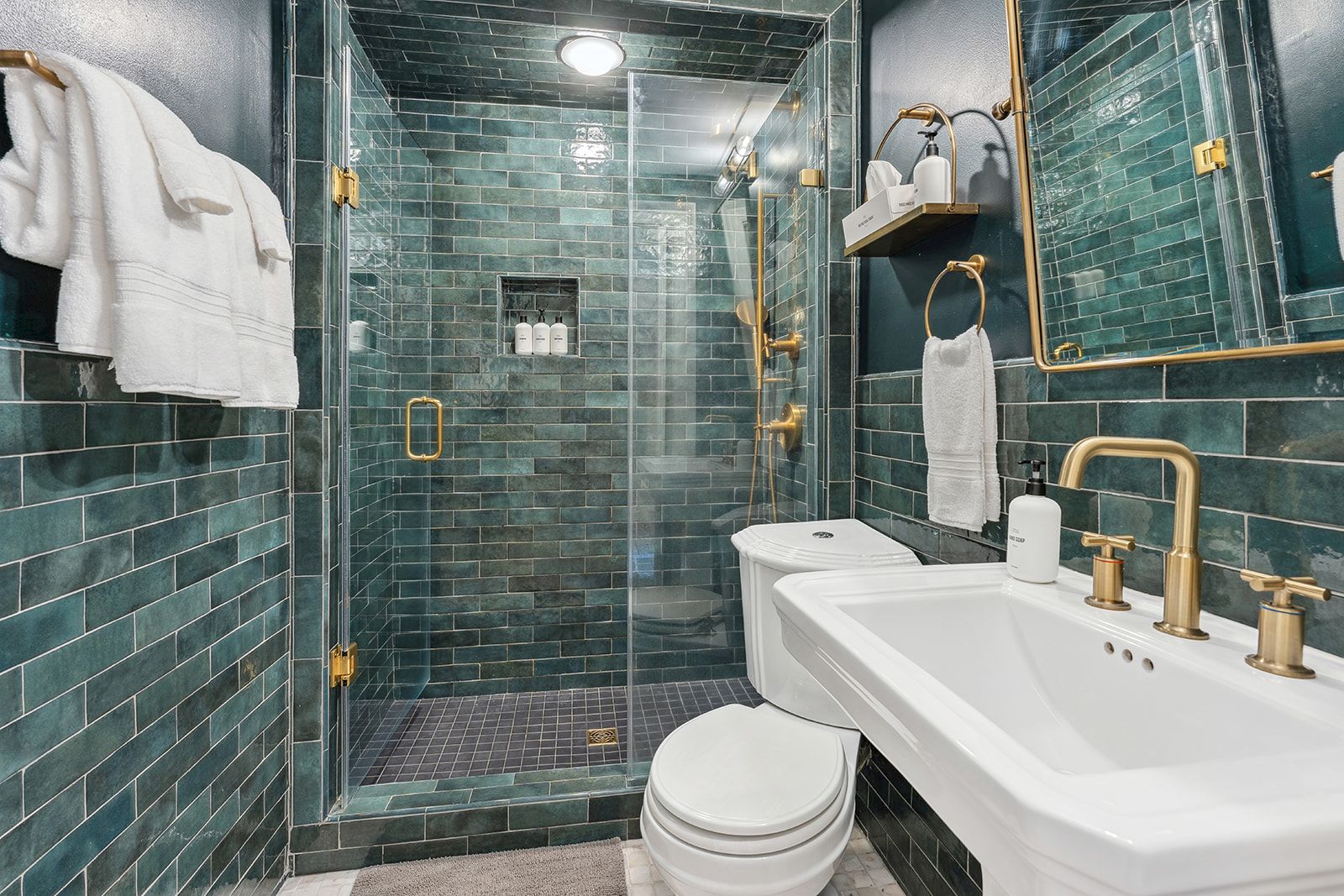 A modern green-tile bathroom featuring a glass shower, gold fixtures, a white sink, and a white toilet. The area looks pristine and coordinated.