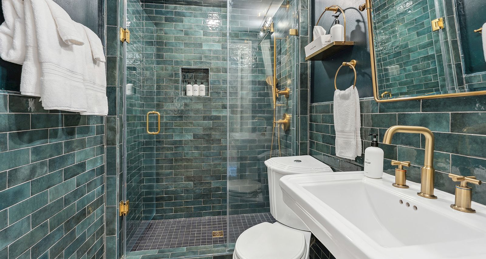 A modern green-tile bathroom featuring a glass shower, gold fixtures, a white sink, and a white toilet. The area looks pristine and coordinated.