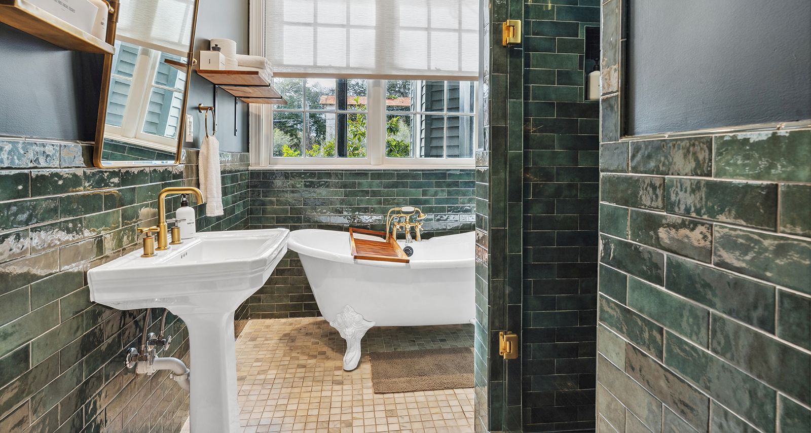 A vintage-style bathroom with green subway tiles, two white pedestal sinks, a freestanding bathtub, a window with natural light, and brass fixtures, all in a compact layout.