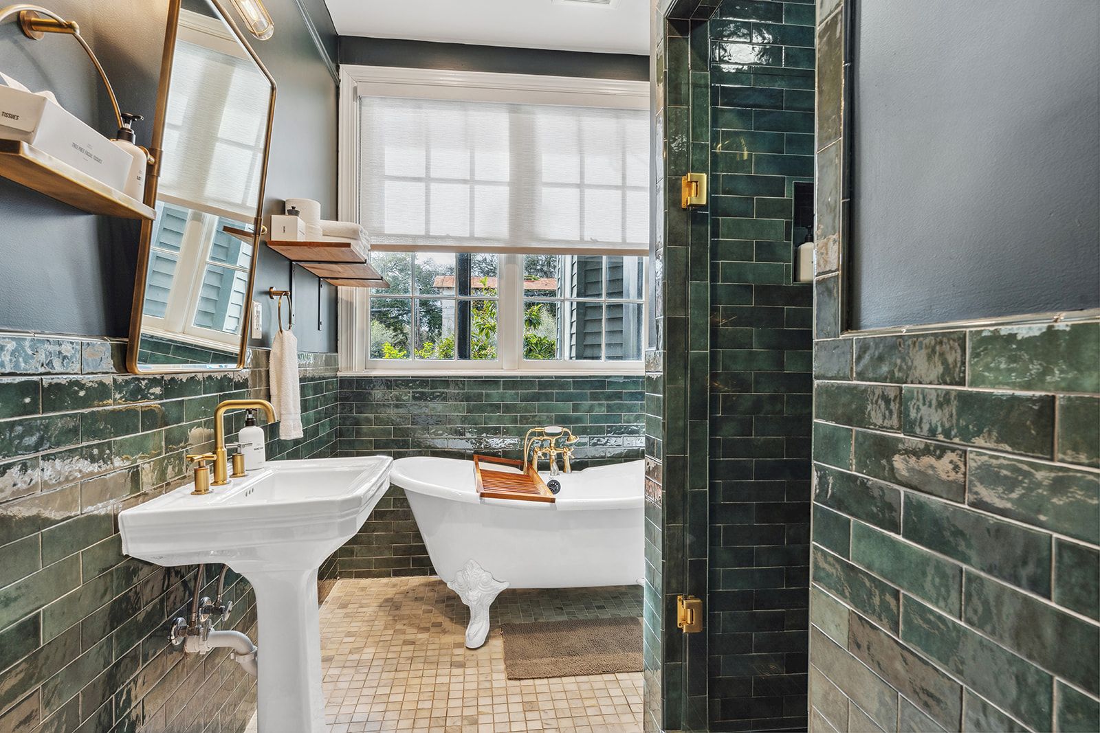 A vintage-style bathroom with green subway tiles, two white pedestal sinks, a freestanding bathtub, a window with natural light, and brass fixtures, all in a compact layout.