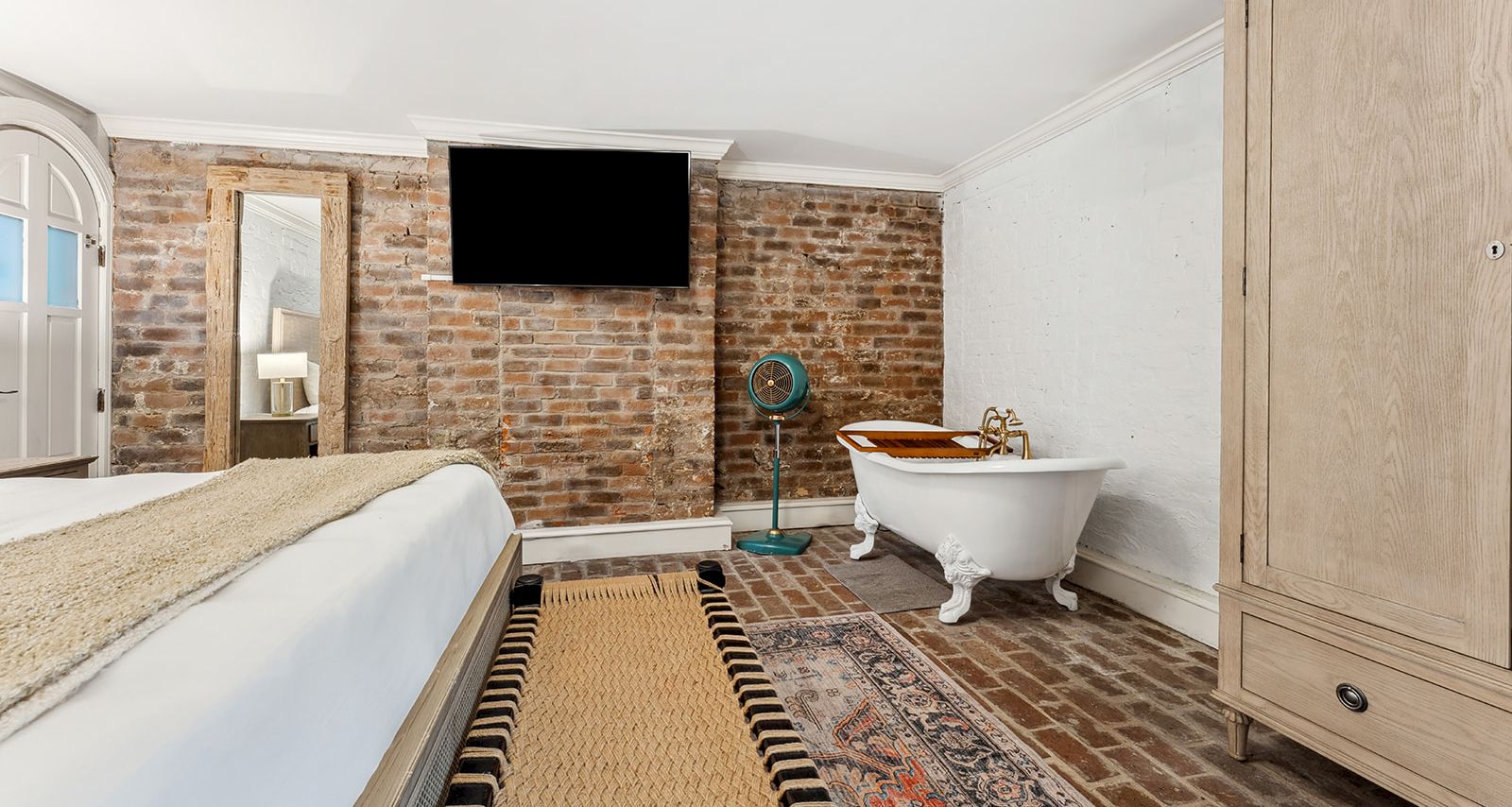 A stylish bedroom with a brick accent wall, mounted TV, a freestanding tub, a tall wooden wardrobe, and a mix of rugs on a brick floor.