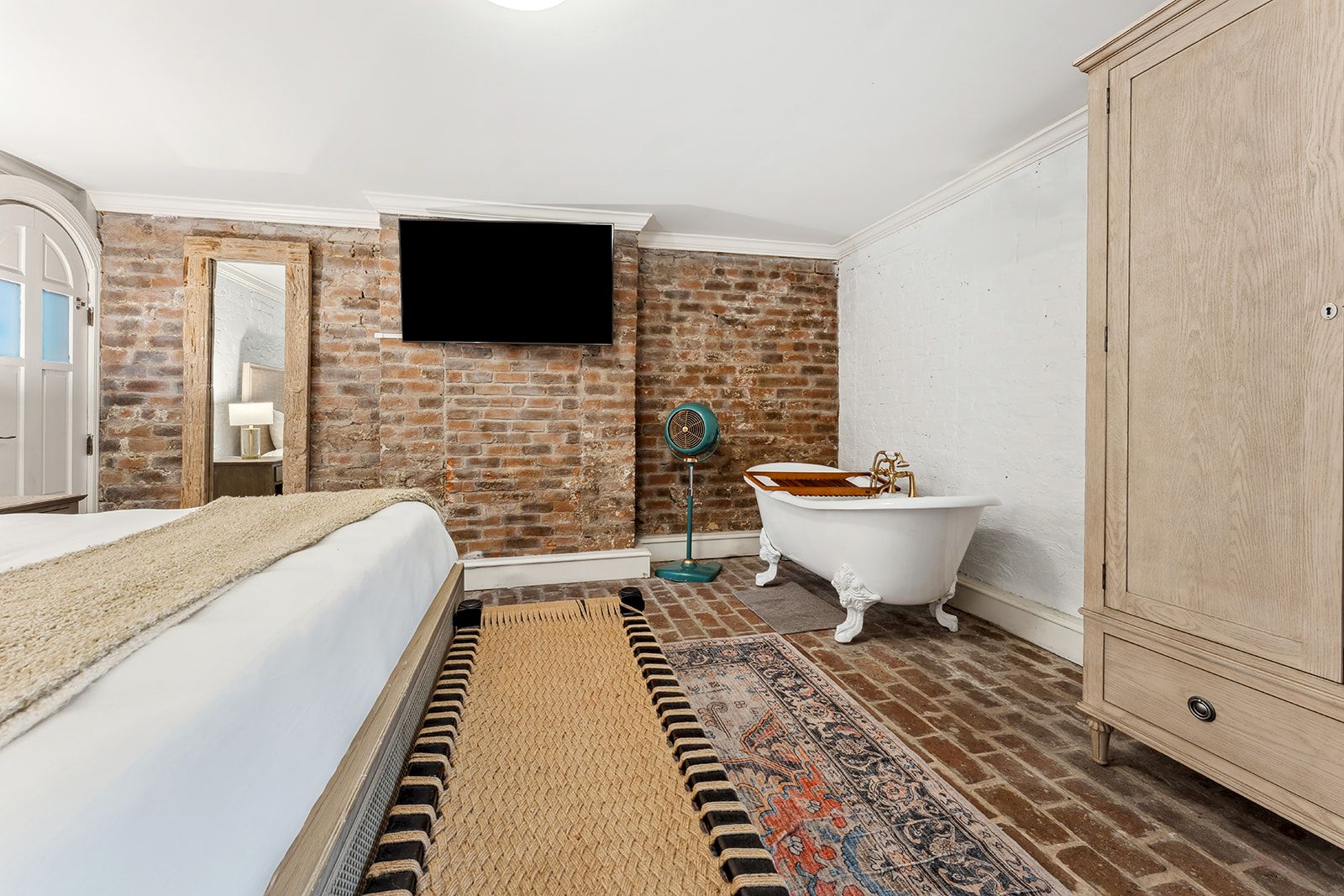 A stylish bedroom with a brick accent wall, mounted TV, a freestanding tub, a tall wooden wardrobe, and a mix of rugs on a brick floor.