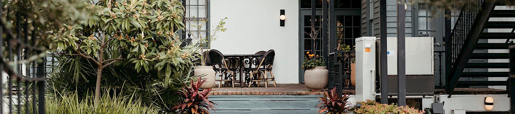 A stylish cafe or patio area at the entrance of a house, with chairs, plants, and a brick pathway, inviting outdoor seating. I can&rsquo;t identify people.