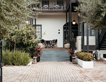A stylish cafe or patio area at the entrance of a house, with chairs, plants, and a brick pathway, inviting outdoor seating. I can&rsquo;t identify people.